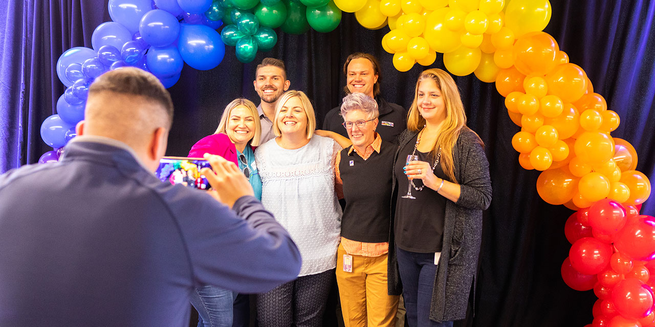 A group of RJF Pro associates pose for a photo under an arch of rainbow colored balloons at a corporate event