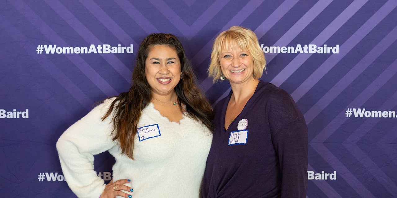 Two RJF Pro associates in front of a purple banner with #womenatbaird hashtags